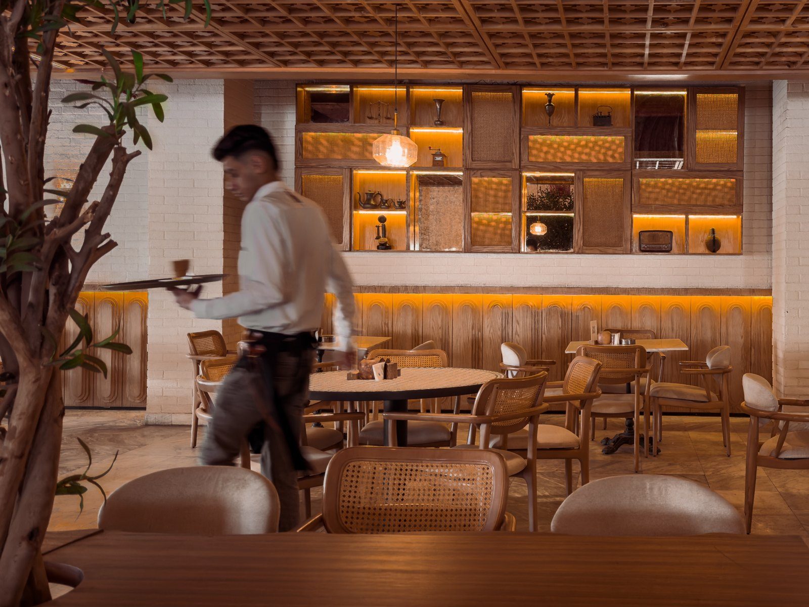 The dining room at Mosaïque: a waiter crossing the floor, cane chairs and a lit wooden shelf wall