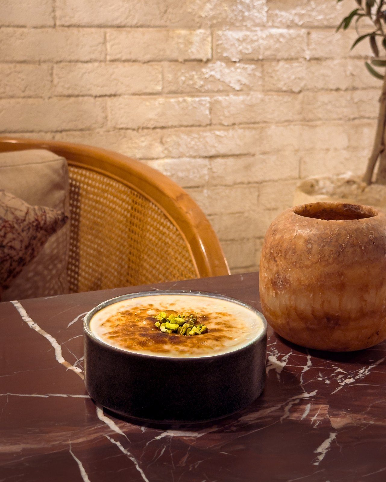 A pistachio-topped dessert in a black bowl on burgundy marble, with a cane chair behind